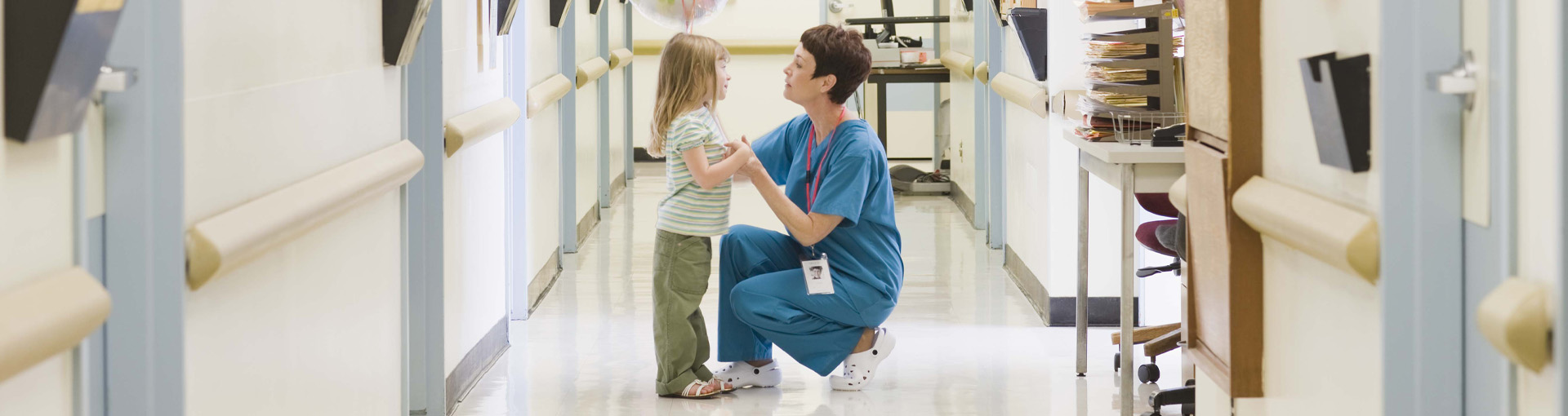 Een zorgprofessional in blauwe scrubs knielt in een ziekenhuisgang en heeft interactie met een jong kind. De zorgprofessional lijkt het kind te troosten of met het kind te praten, dat staat en naar hen kijkt. De gang is goed verlicht en omgeven door medische apparatuur en deuren. De merknaam DNV is geassocieerd met deze afbeelding, wat duidt op een focus op kwaliteitszorgdiensten of accreditatie.