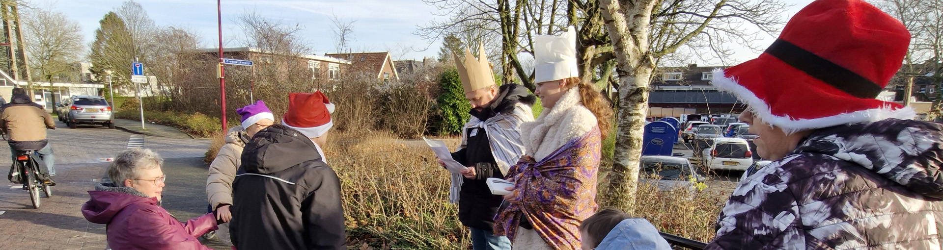 Groep mensen met feestelijke hoeden en winterkleding buiten in een woonwijk tijdens het feestseizoen, met DNV