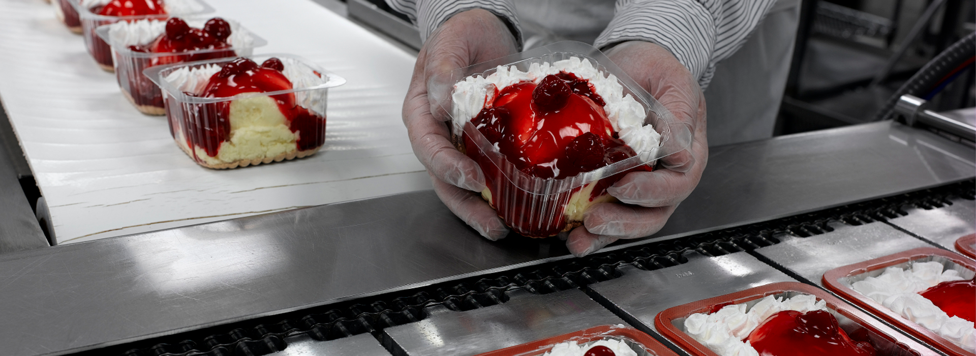 Persoon houdt een dessertbeker met lagen cake, slagroom en aardbeien vast in een voedselproductielijn, met identieke dessertbekers op een lopende band op de achtergrond.
