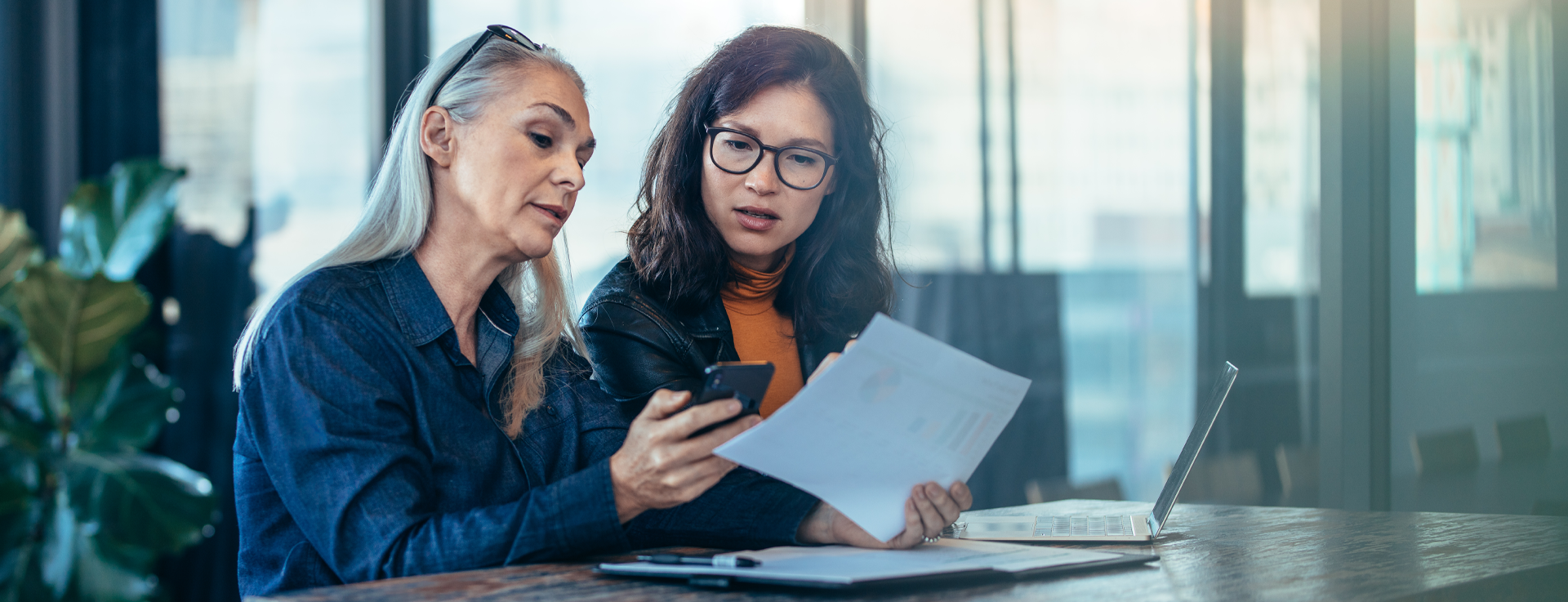 Twee personen zitten aan een tafel in een kantooromgeving, waarbij één persoon een papier vasthoudt en de ander een pen, met een laptop op tafel.
