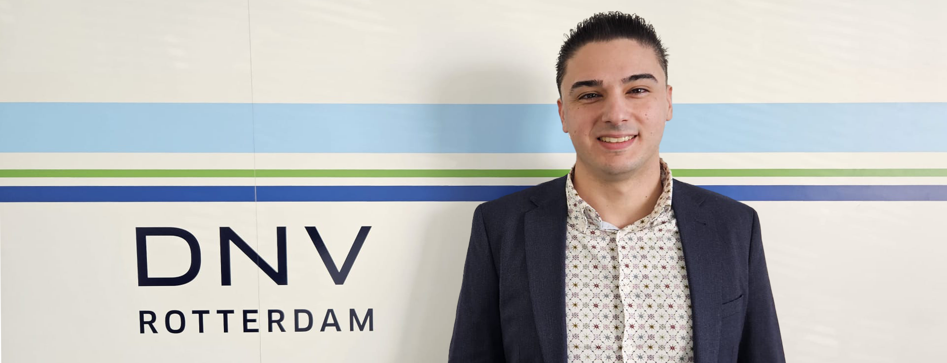 Een persoon die voor een muur staat met de tekst "DNV ROTTERDAM", gekleed in een donkere blazer en lichtgekleurde shirt.