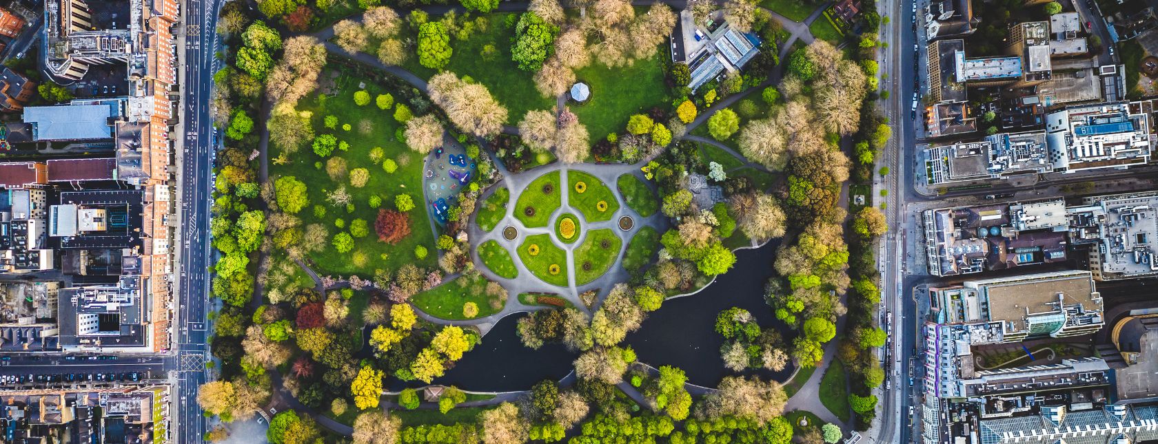 Een luchtfoto van een groot, rechthoekig park omringd door stedelijke gebouwen, met geometrische paden en vijvers.