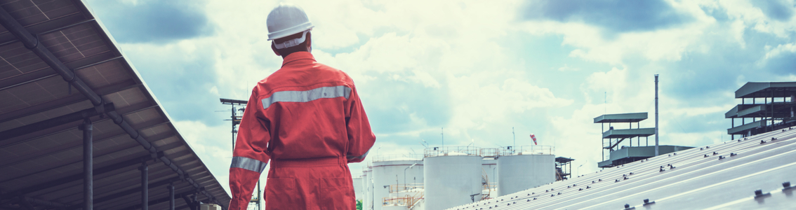 Persoon in rode uniform en witte helm staat in een industriële omgeving met grote tanks en platforms, onder een deels bewolkte hemel.